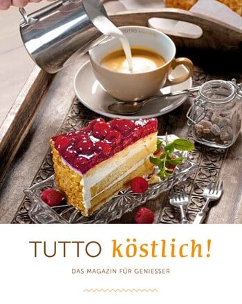 ein Tablett mit einem Stück Himbeertorte und einem Cappucchino mit Milchschaumkanne, dazu ein Glas mit Kandiszucker